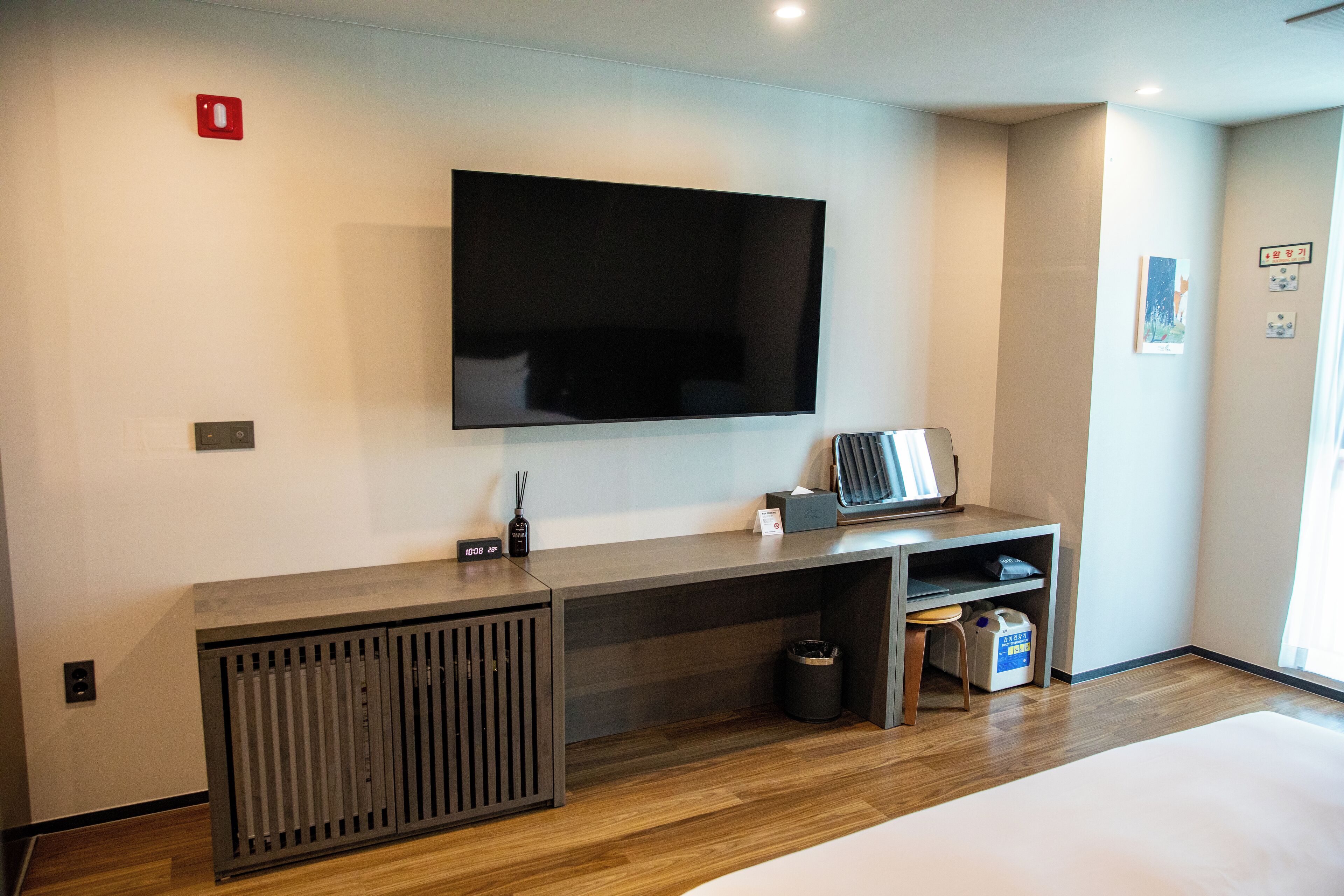 Una habitación de hotel moderna con una amplia televisión de pantalla plana montada en la pared, un escritorio de madera con un cesto de basura y un taburete pequeño.
