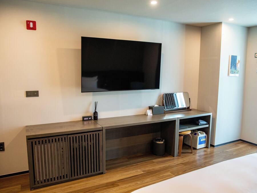 Una habitación de hotel moderna con una amplia televisión de pantalla plana montada en la pared, un escritorio de madera con un cesto de basura y un taburete pequeño.