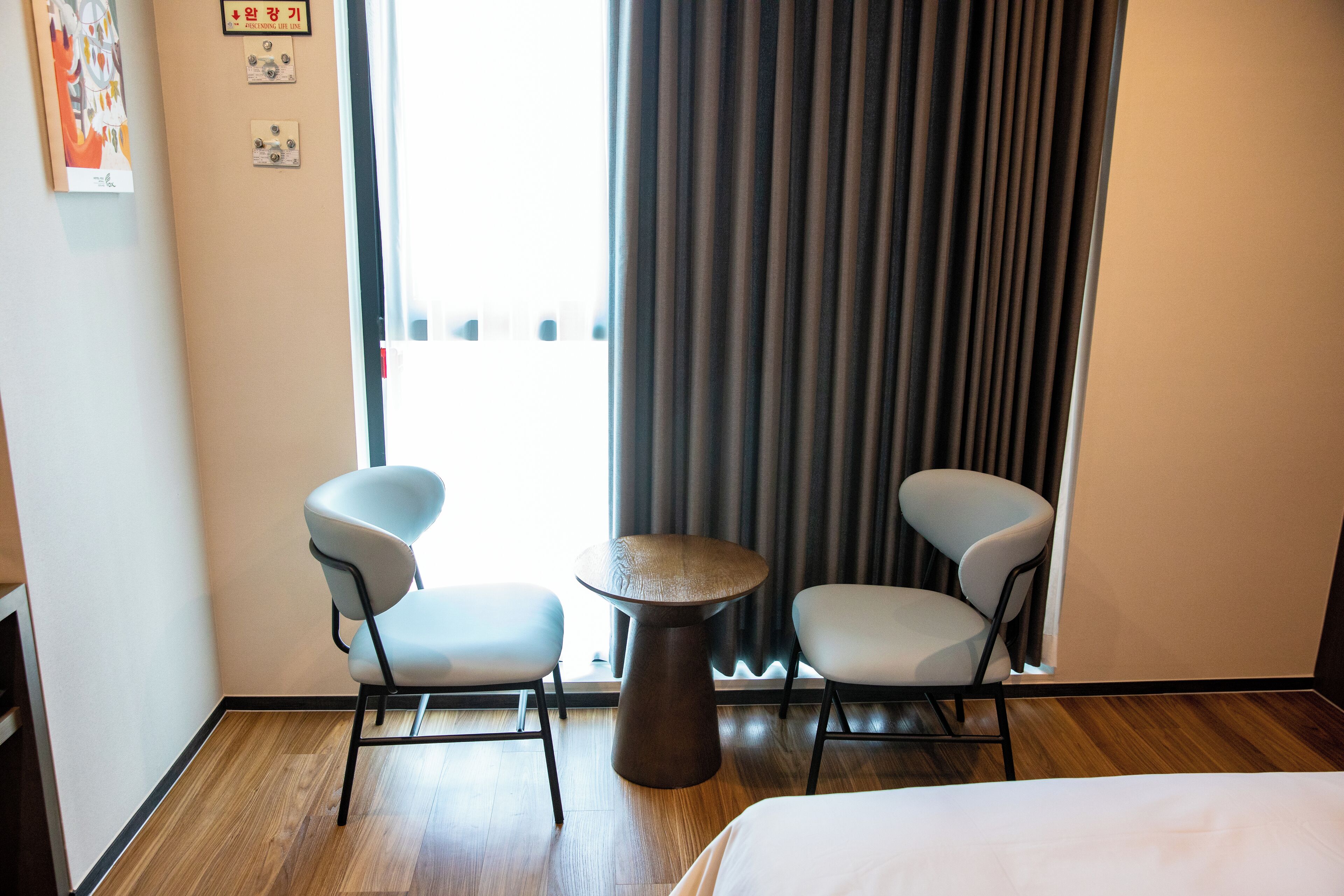Ein Hotelzimmer mit einem Bett, zwei Stühlen, einem kleinen runden Tisch und Blick ins Freie durch ein Vorhangfenster.