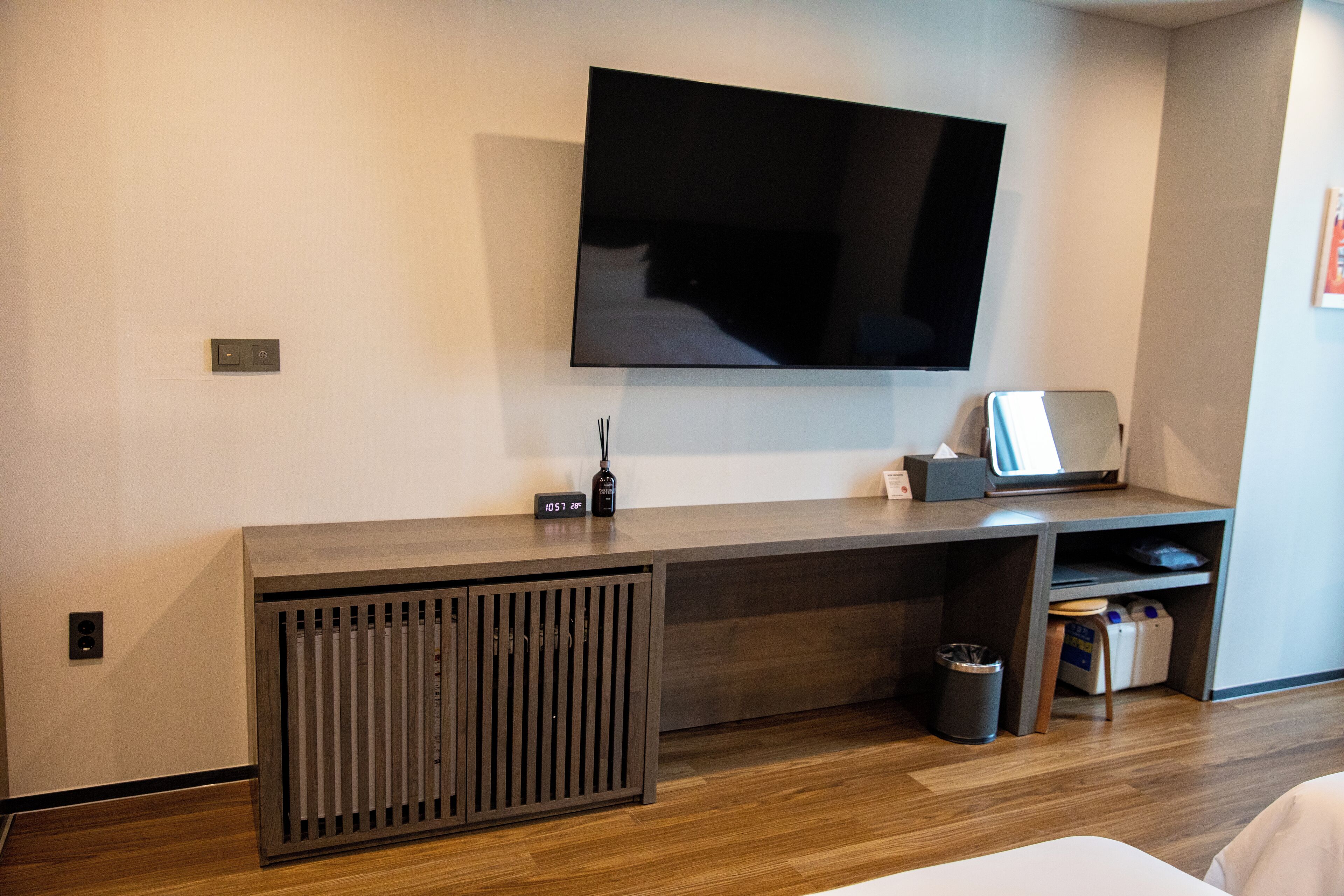A modern hotel room with a large flat-screen TV mounted on the wall, a wooden desk with a laptop, a trash bin, and a small seating area.
