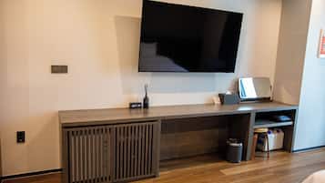 A modern hotel room with a large flat-screen TV mounted on the wall, a wooden desk with a laptop, a trash bin, and a small seating area.