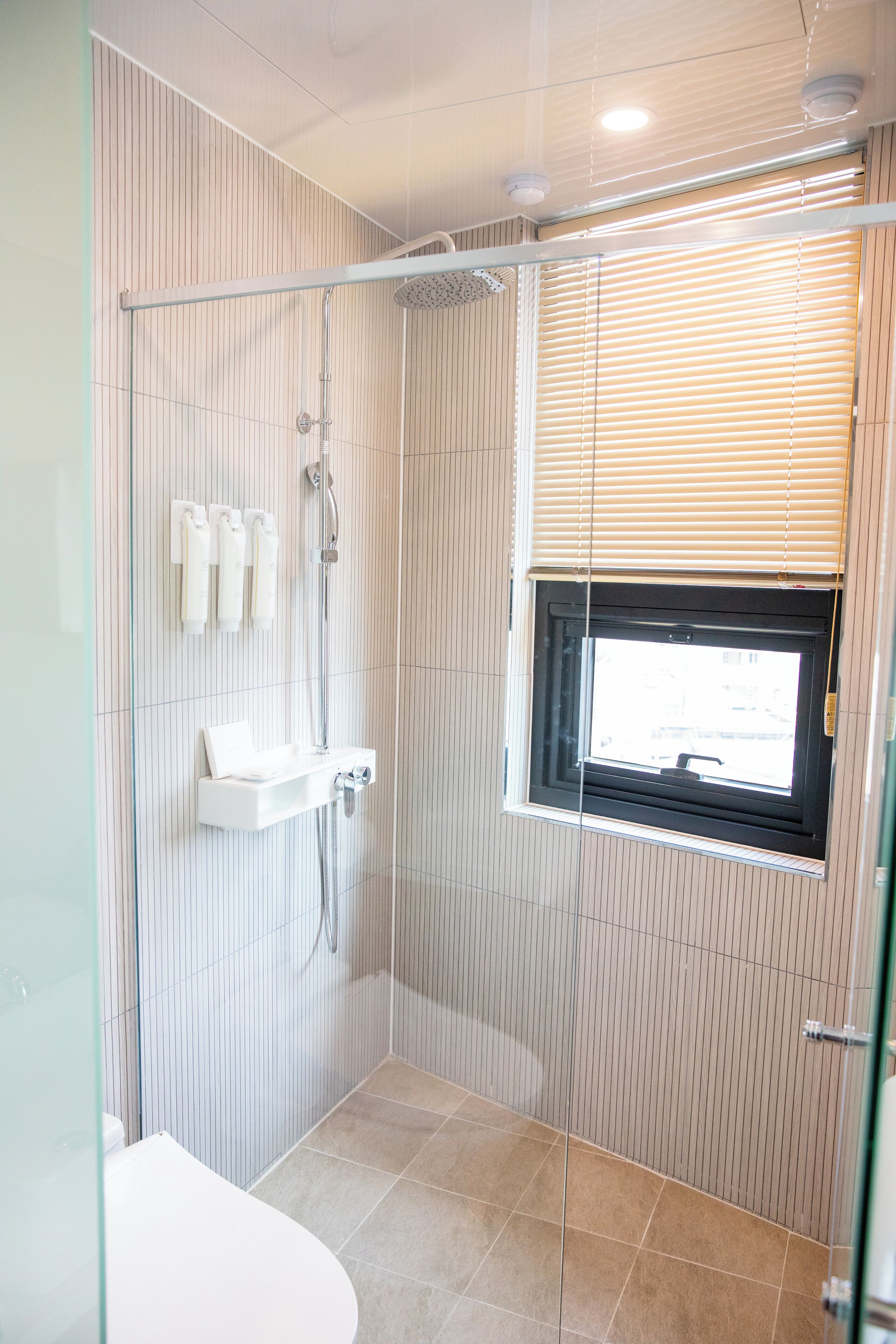 A modern shower with a glass door, a window with blinds, and a white sink with a towel rack.