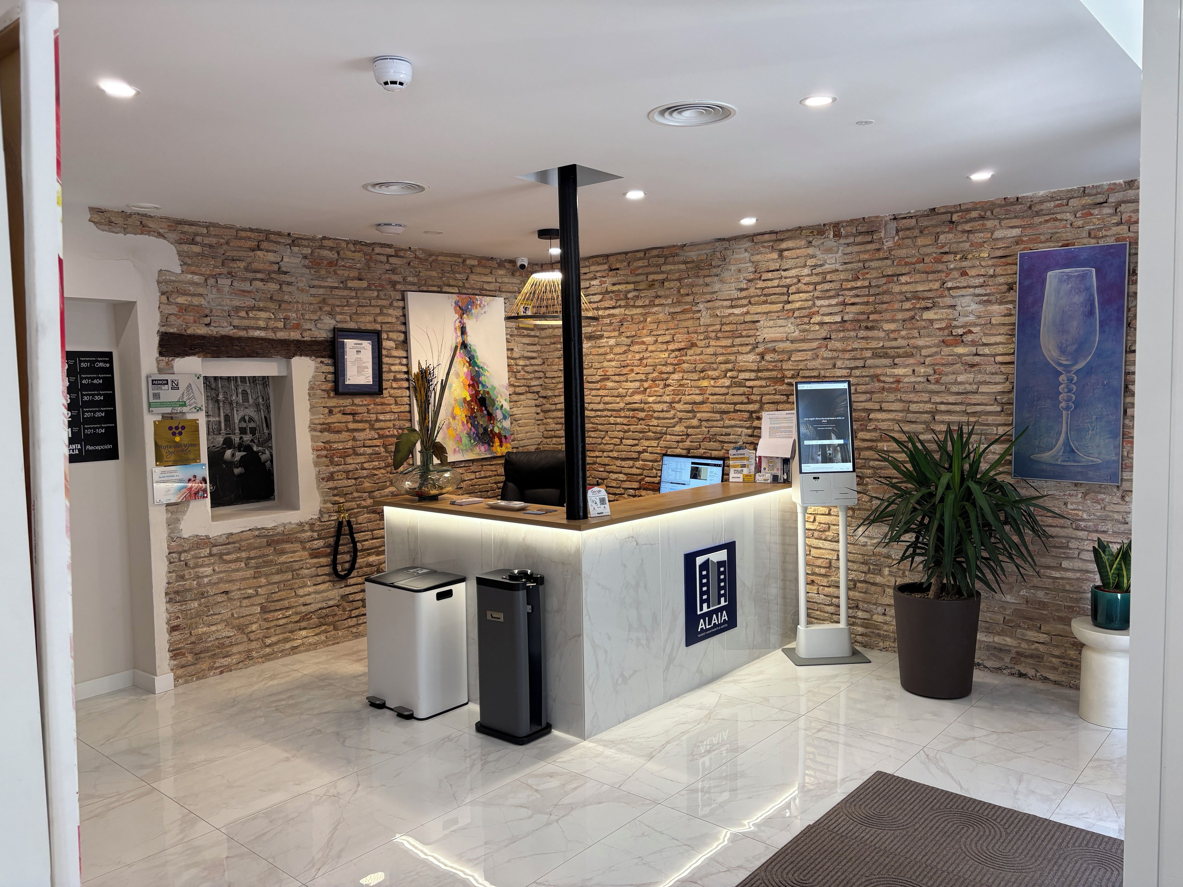 A reception area with a brick wall, a reception desk, a computer, a trash bin, and a potted plant.