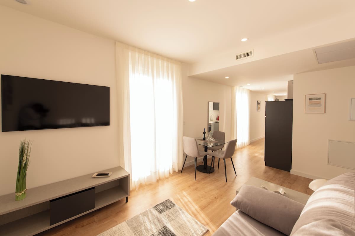 A modern hotel room with a flat-screen TV, a dining area with a table and chairs, a bed, and a large window with curtains.