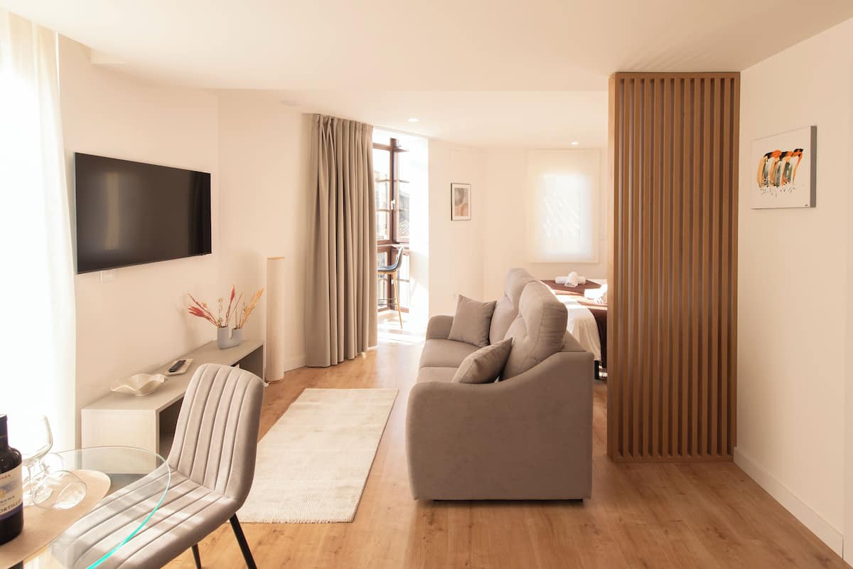A modern living room with a sofa, a TV mounted on the wall, a dining table with chairs, and a wooden floor.