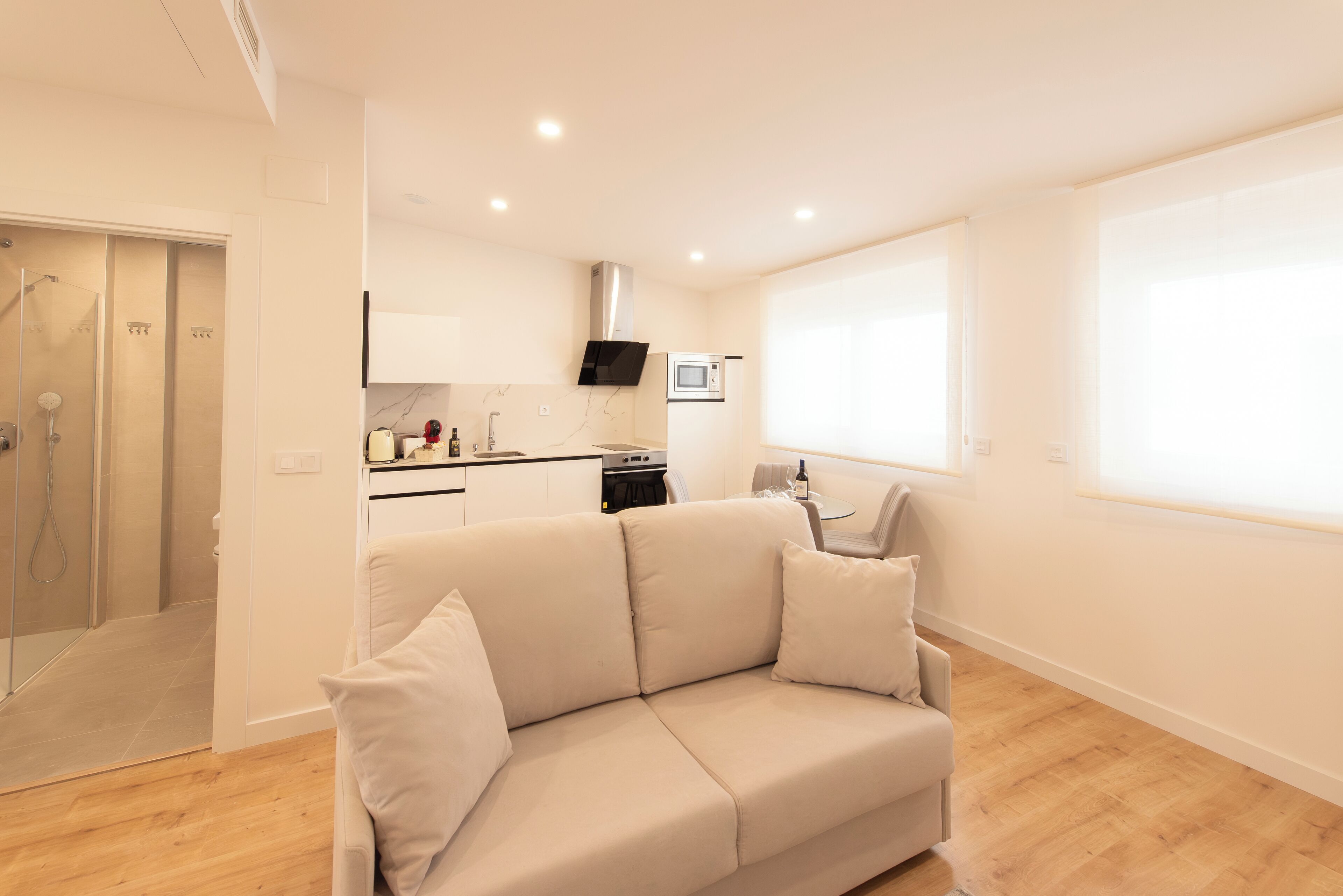 Een modern appartement met een woonruimte, inclusief een beige bank, een keuken met een magnetron en een douchecabine van glas.