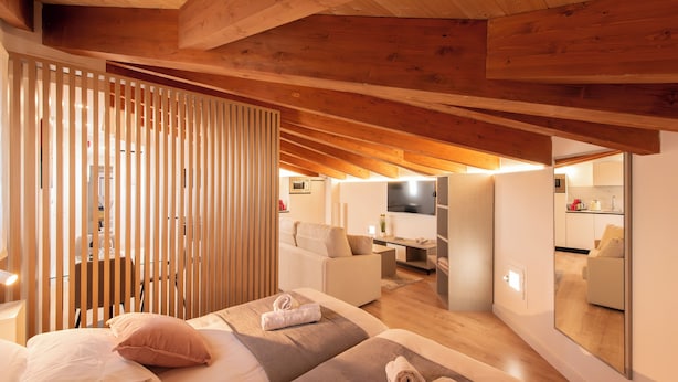 A modern bedroom with a sloped ceiling, wooden beams, a large bed with pillows, a bedside table, and a mirror reflecting the room.