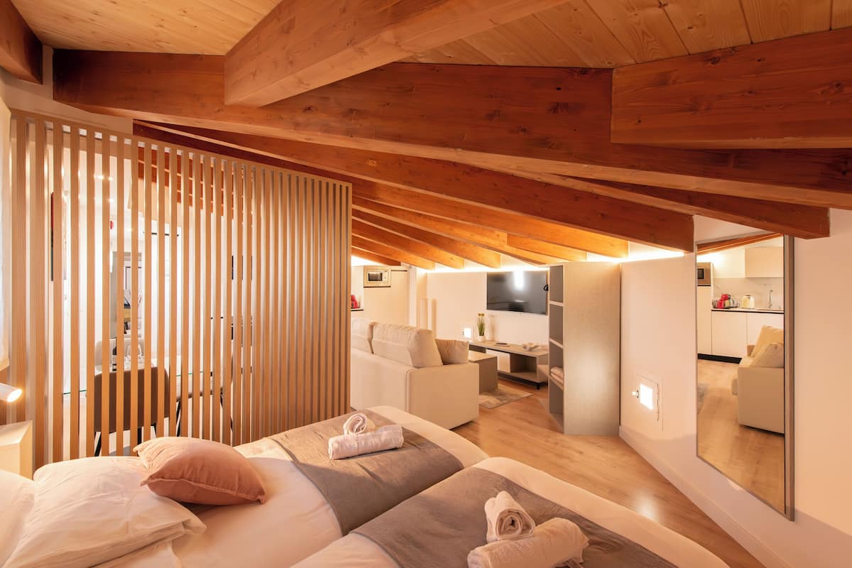 A modern bedroom with a sloped ceiling, wooden beams, a large bed with pillows, a bedside table, and a mirror reflecting the room.