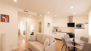 A modern living room with a beige sofa, a glass dining table, and a kitchen area with a microwave and stove.