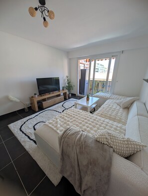 A modern bedroom with a large bed, a TV, a small table, and a balcony with a view of buildings. - T3 near train station - air conditioning, 15 min walk to beaches (CAGNES SUR MER)