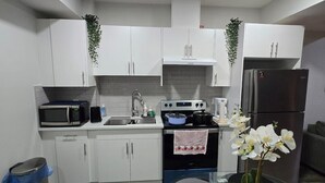 A modern kitchen with white cabinets, a microwave, stove, and refrigerator. There is a dining table with a vase of flowers. - Stylish 2BD Basement Suite Close to WEM (Edmonton)