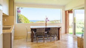 Ein Essbereich mit großem Fenster und Blick auf eine reizvolle Landschaft, einem Tisch mit Korbstühlen und einer Blumenvase.