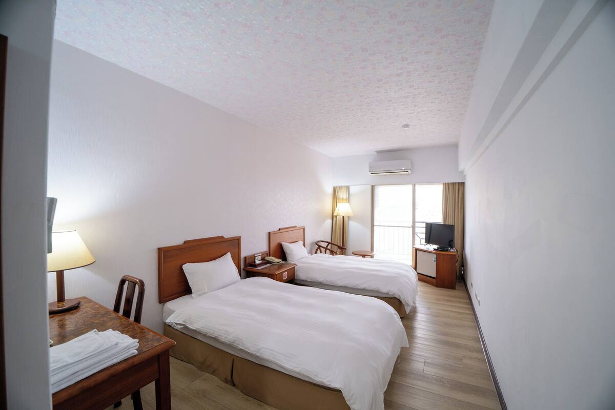 A hotel room with two beds, a desk with a lamp, a chair, a TV, and a window with curtains.