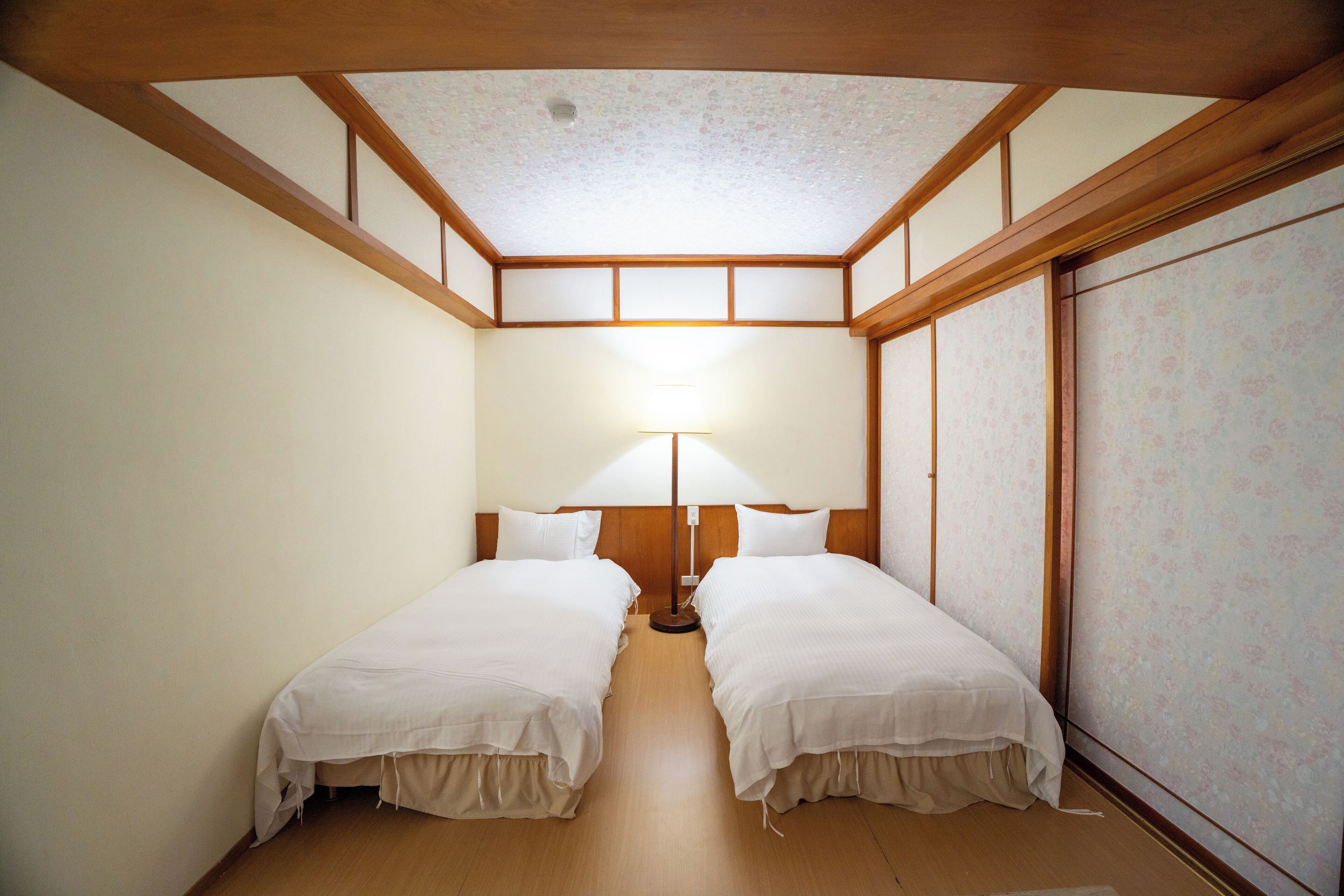 Une chambre avec deux lits, des poutres en bois au plafond, une grande fenêtre ornée d’un papier peint à motifs floraux et une table de chevet avec une lampe.
