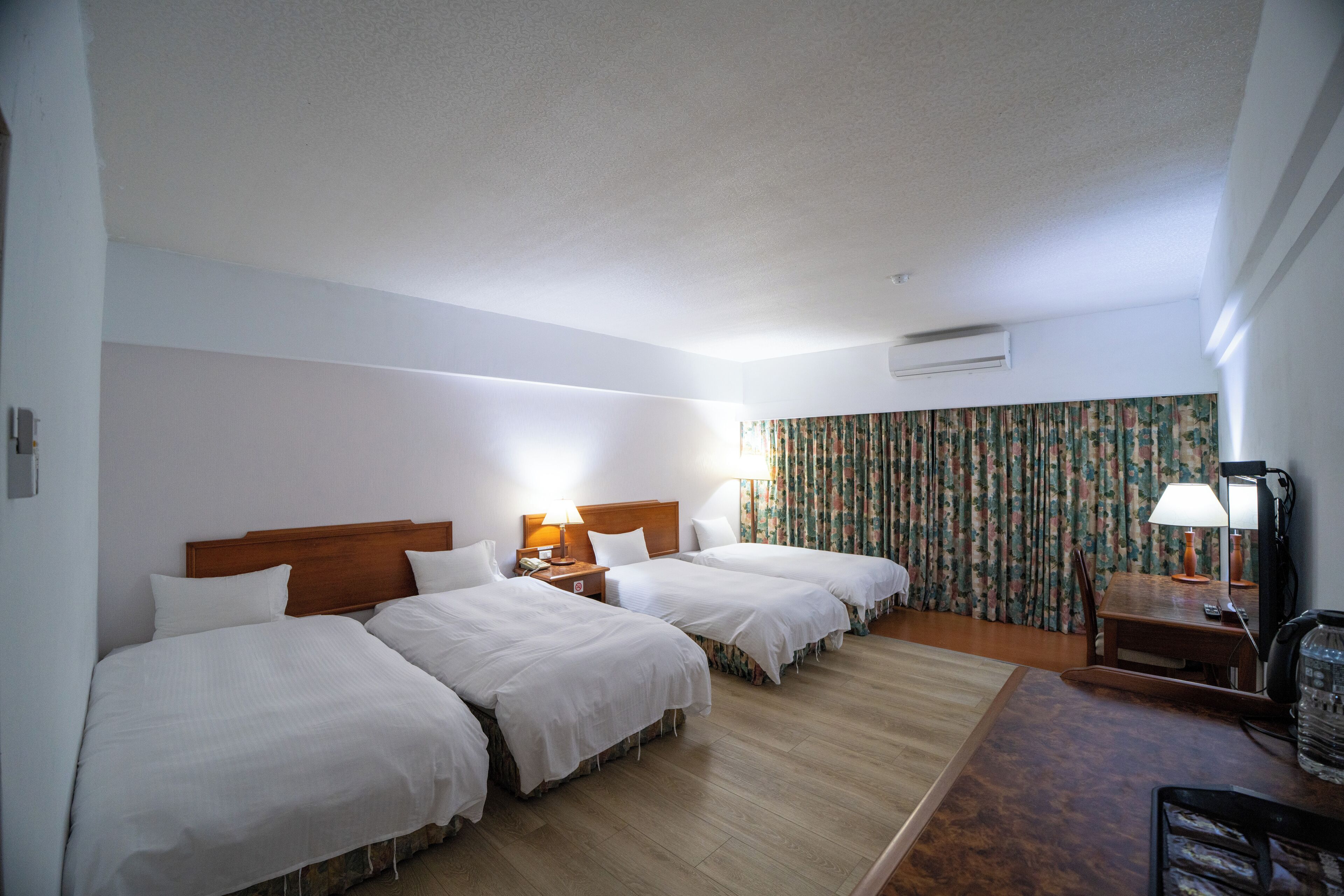 A hotel room with three beds, a wooden headboard, a desk with a lamp, and a floral curtain.