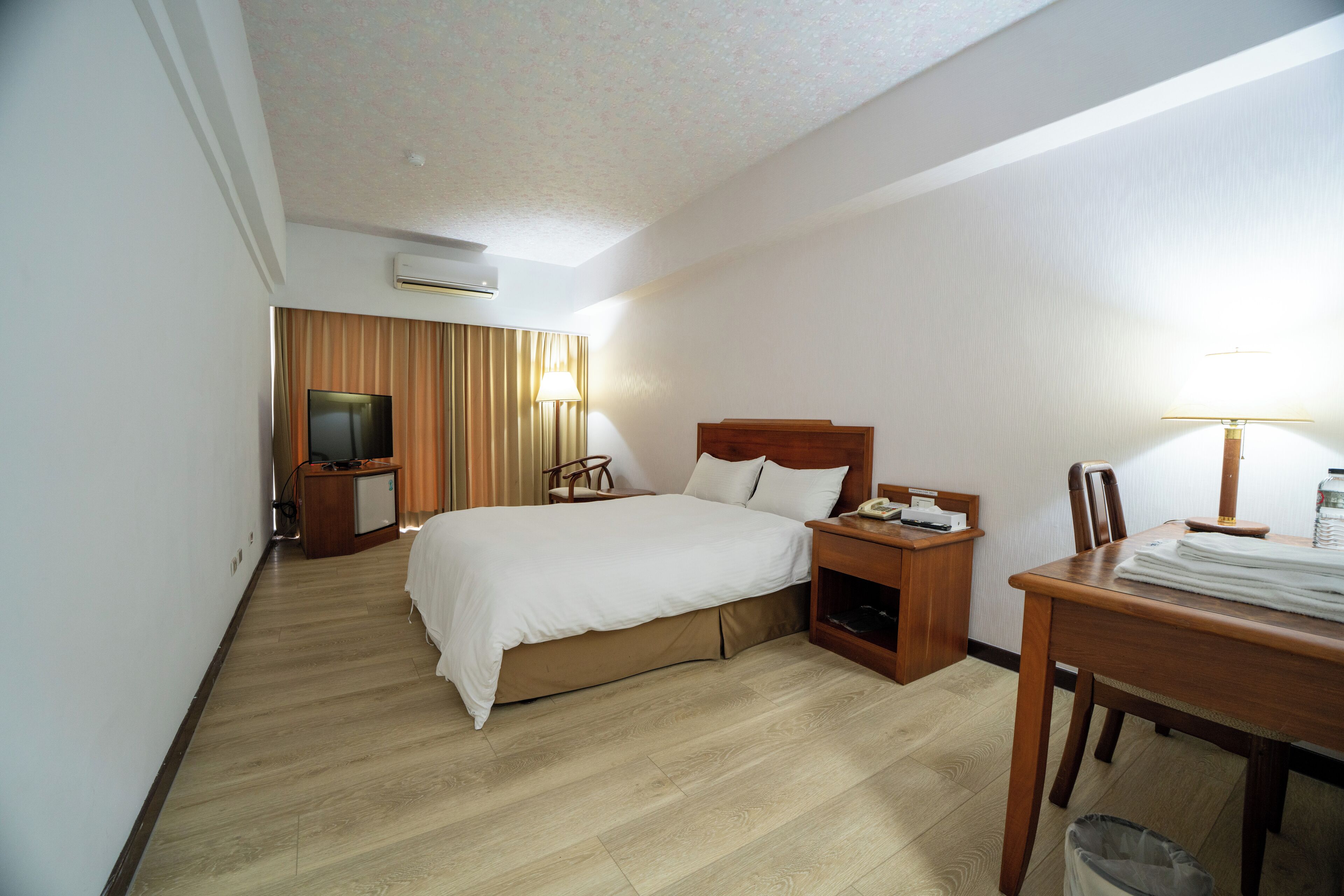 A hotel room with a bed, bedside table, lamp, TV, and a desk with a chair.