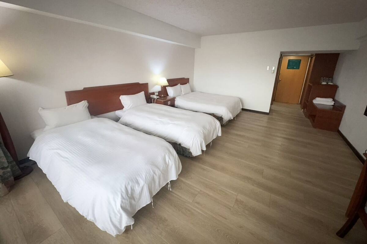 A hotel room with two single beds, a wooden headboard, a bedside table with a lamp, and a wooden floor.