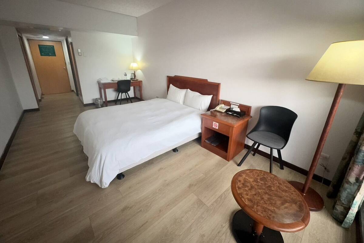 A hotel room with a bed, a desk, a chair, a small table, and a lamp.