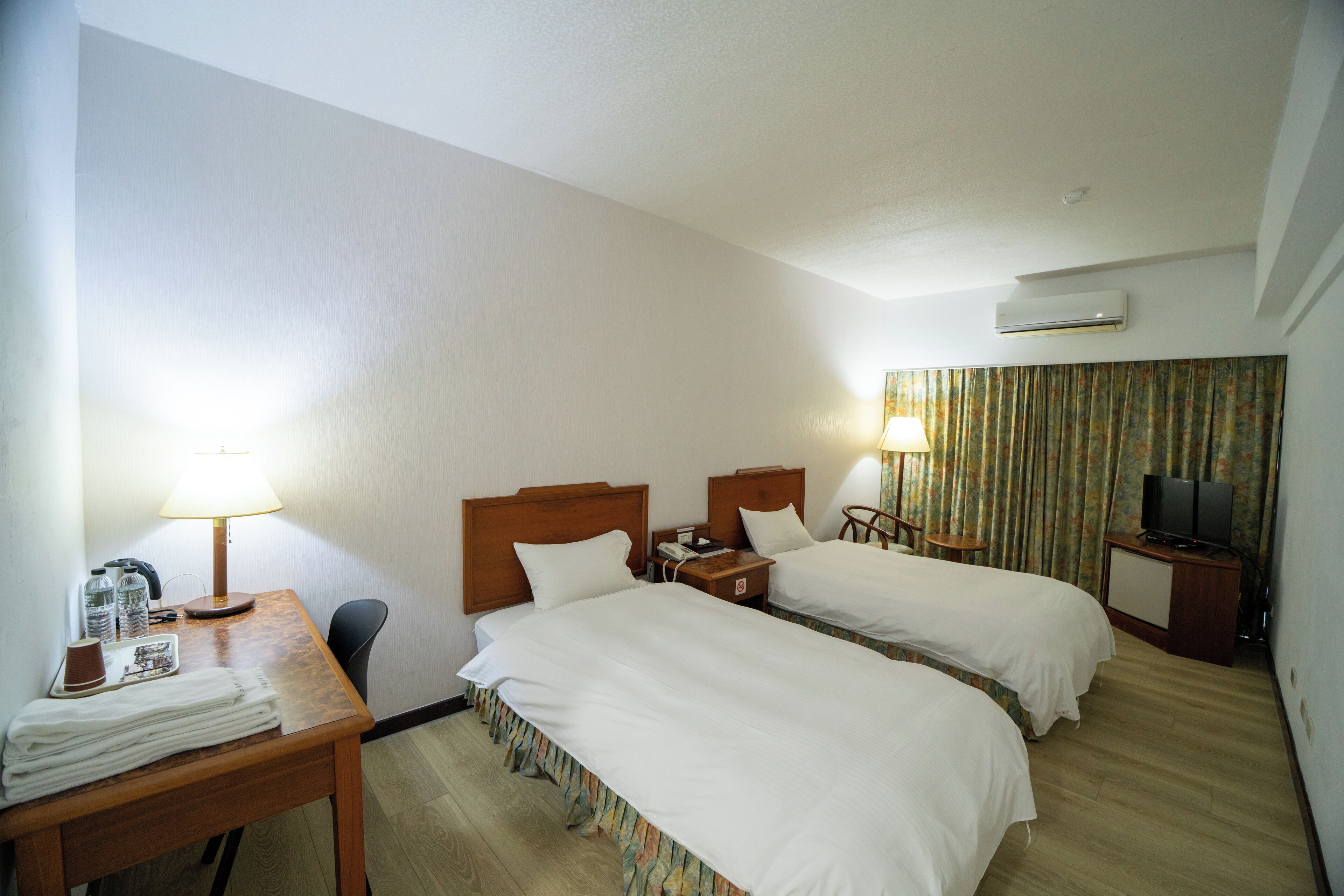 A hotel room with two beds, a desk with a lamp, a telephone, a television, and an air conditioning unit.