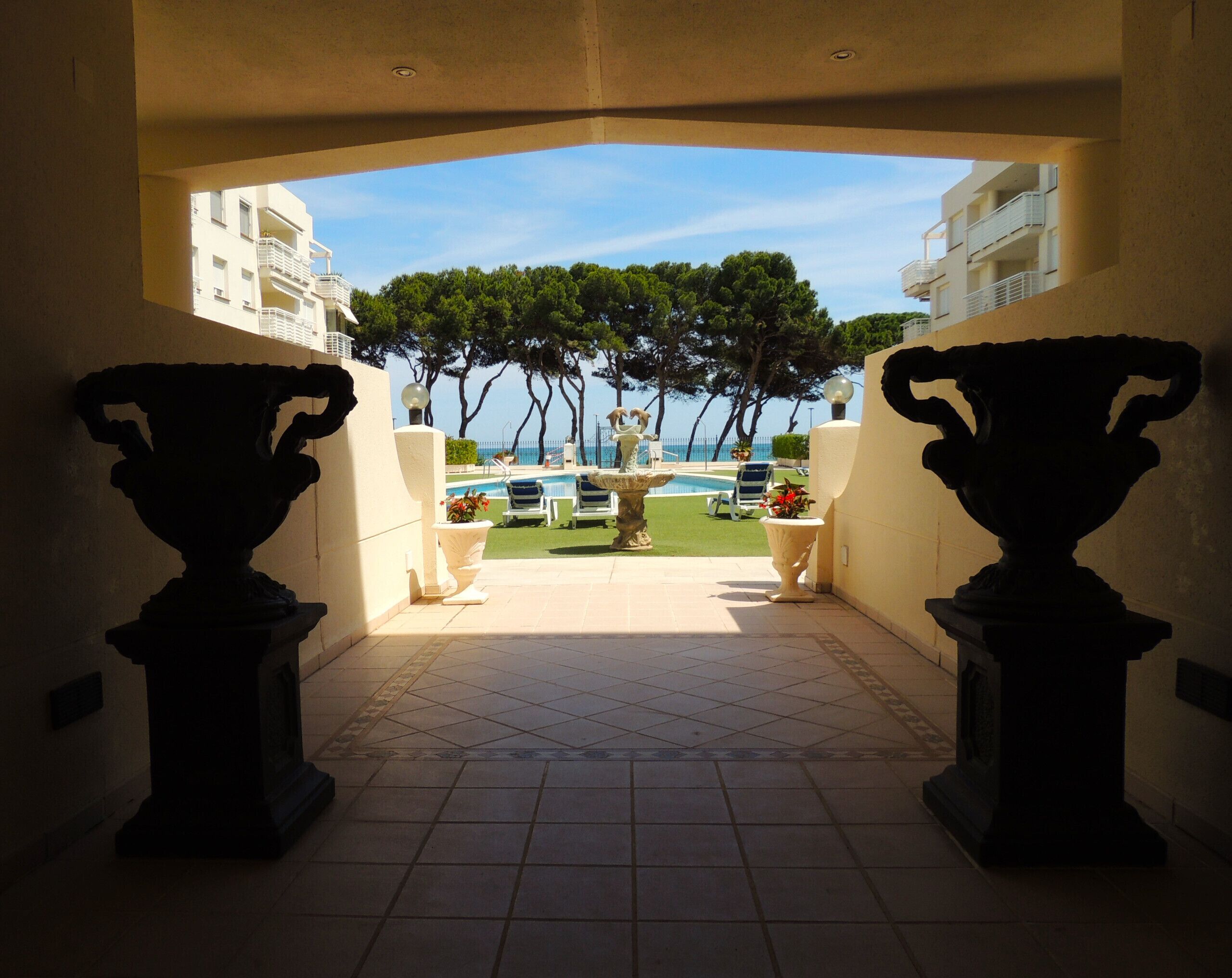 A view through a corridor with two large vases on pedestals, leading to a pool area with a clear sky and trees in the background.