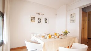 Een eetruimte met een tafel voor vier personen, witte stoelen, een raam met gordijnen en ingelijste foto's aan de muur.
