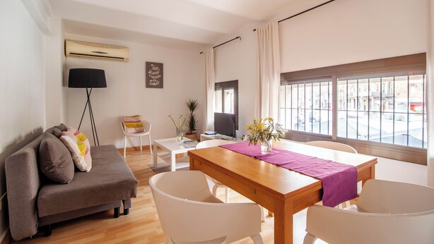 Een moderne woonkamer met een bank, eettafel en stoelen. Er is een televisie, een plant en een raam met uitzicht op de straat.