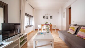 Een moderne woonkamer met een flatscreen televisie, een witte salontafel, een bank met gestreepte kussens en een eethoek met een houten tafel en stoelen.