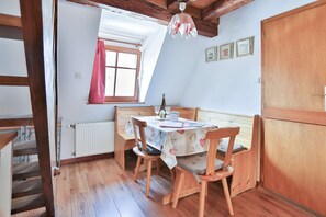 A dining area with a table set for two, wooden chairs, a chandelier, and a window with a red curtain.