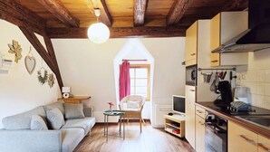 Una sala de estar acogedora con un sofá gris, una mesita pequeña y una silla de mimbre. Se ve una cocina con estufa, microondas y armarios.