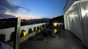 Una terraza en la azotea con mesa y sillas, vista panorámica de la ciudad y un edificio blanco al fondo.