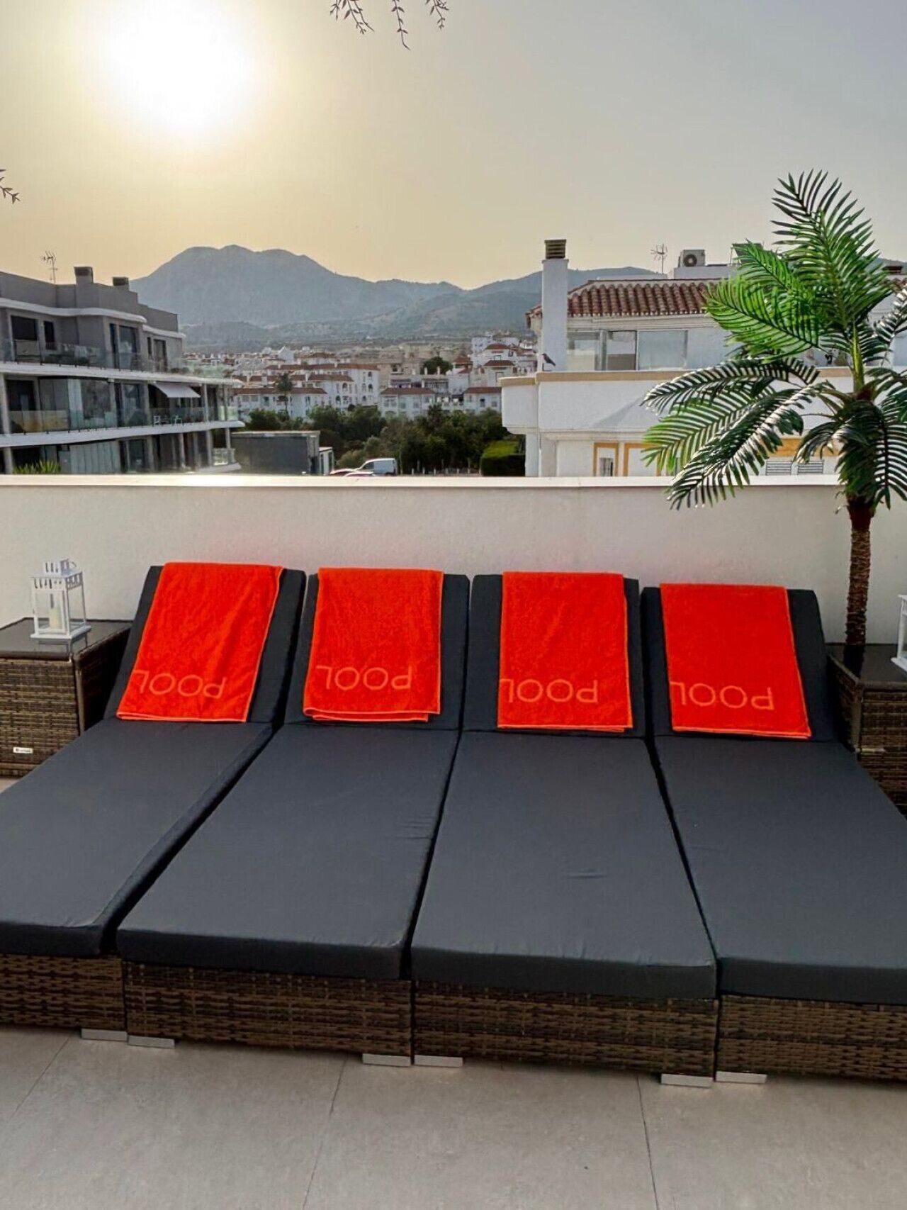 Eine Dachterrasse mit vier schwarzen Liegestühlen, jeder mit einem roten Handtuch und dem Schriftzug 'POOL'. Eine Palme ist im rechten Bildbereich sichtbar.