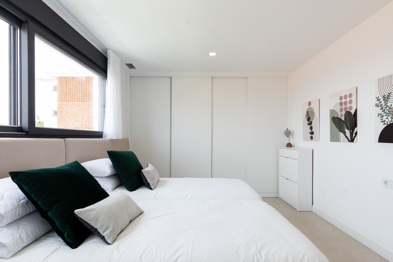 Quarto moderno com uma cama grande, roupa de cama branca e almofadas verdes. Há uma janela com vista para um edifício.