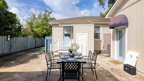 A patio with a table set for four, surrounded by chairs, and a small house in the background.