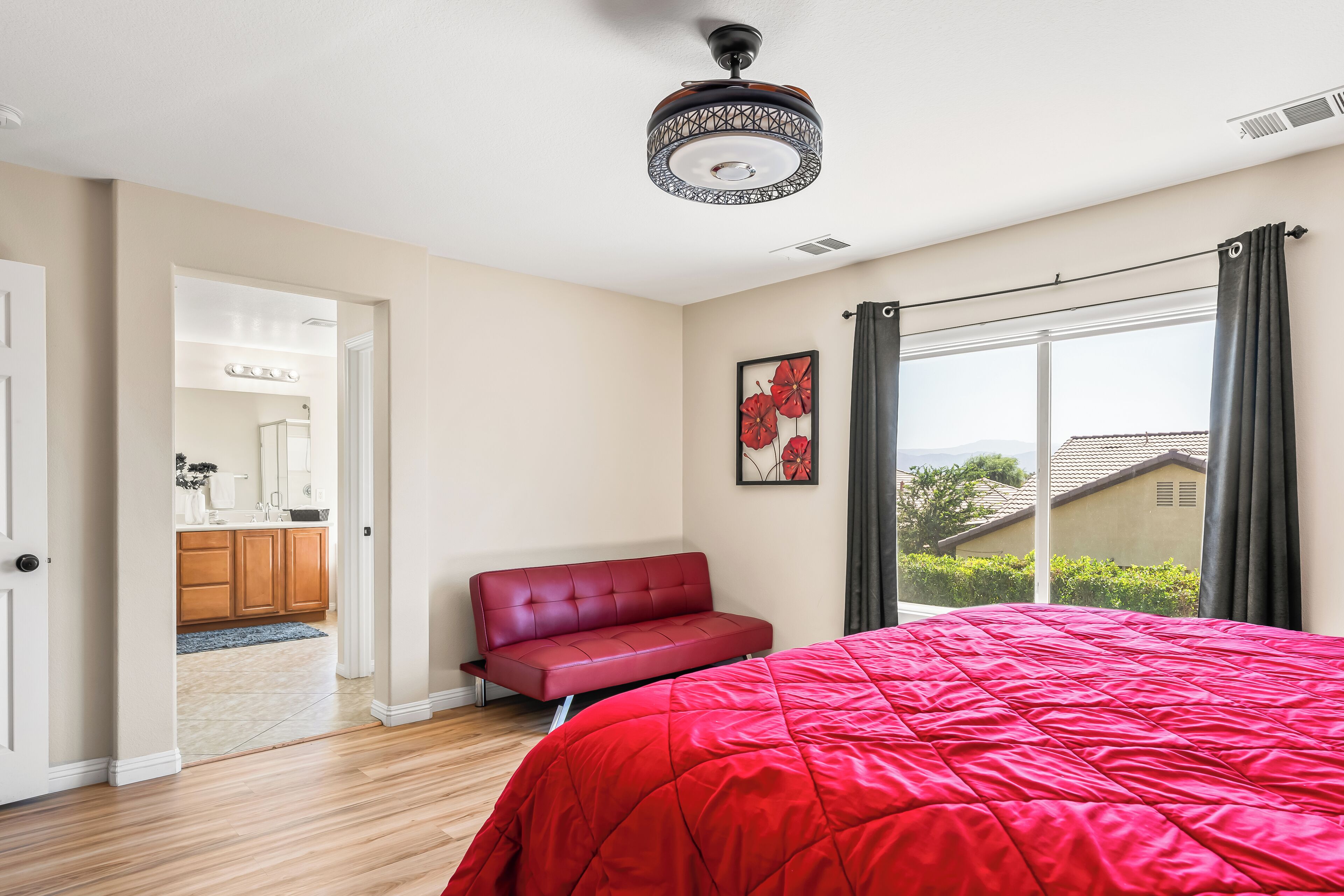 Ein Schlafzimmer mit roter Bettdecke, einem roten Sofa, einem Deckenventilator, einem Fenster mit Vorhängen und Blick auf ein Haus.