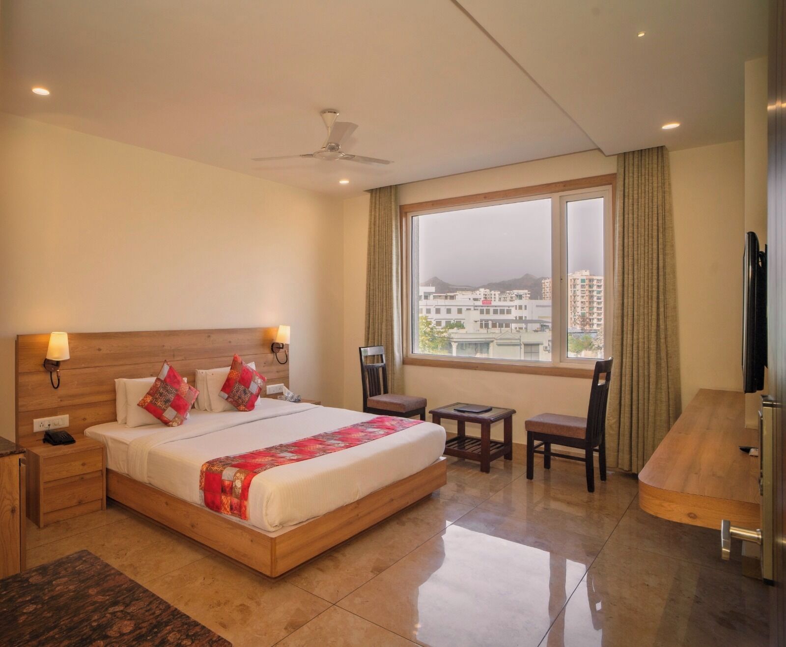 A hotel room with a bed, bedside tables, a desk, a chair, a TV, and a large window with a city view.