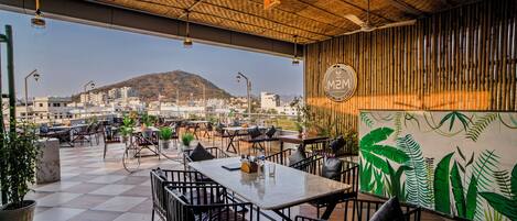 An outdoor café with tables and chairs, a view of a cityscape, and a bamboo wall.