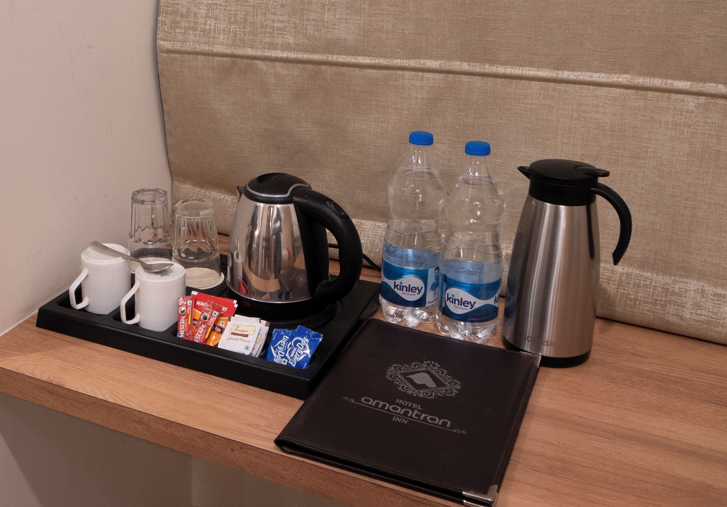Minibar de hotel com uma bandeja contendo duas canecas brancas, um copo, uma chaleira, duas garrafas de água Kinley, uma cafeteira de aço inoxidável e um livreto preto com a inscrição 