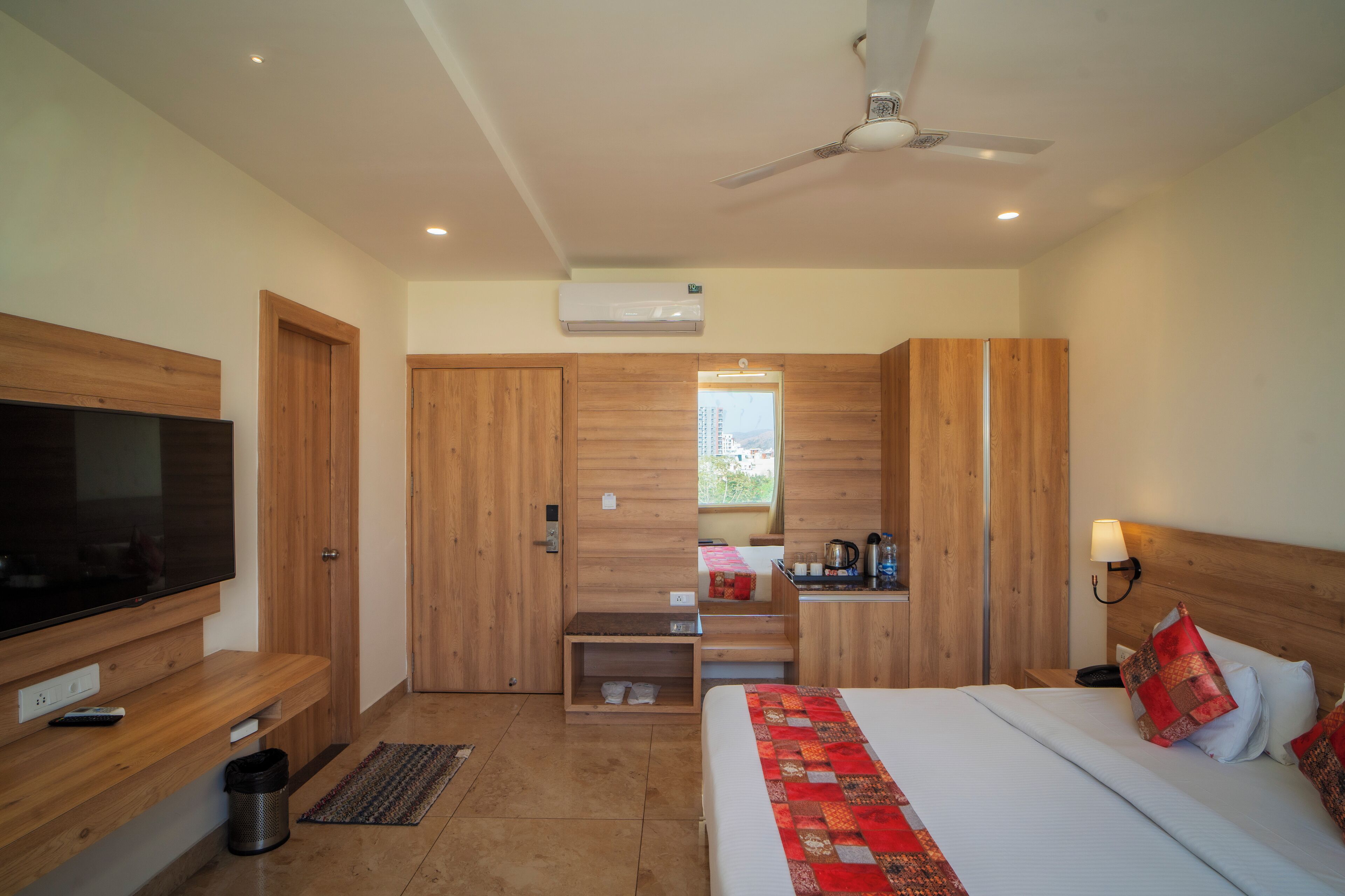 Un dormitorio moderno con una televisión de pantalla plana, una cama con colcha estampada en rojo y blanco, un armario de madera, una pequeña cocina americana y un ventilador de techo.
