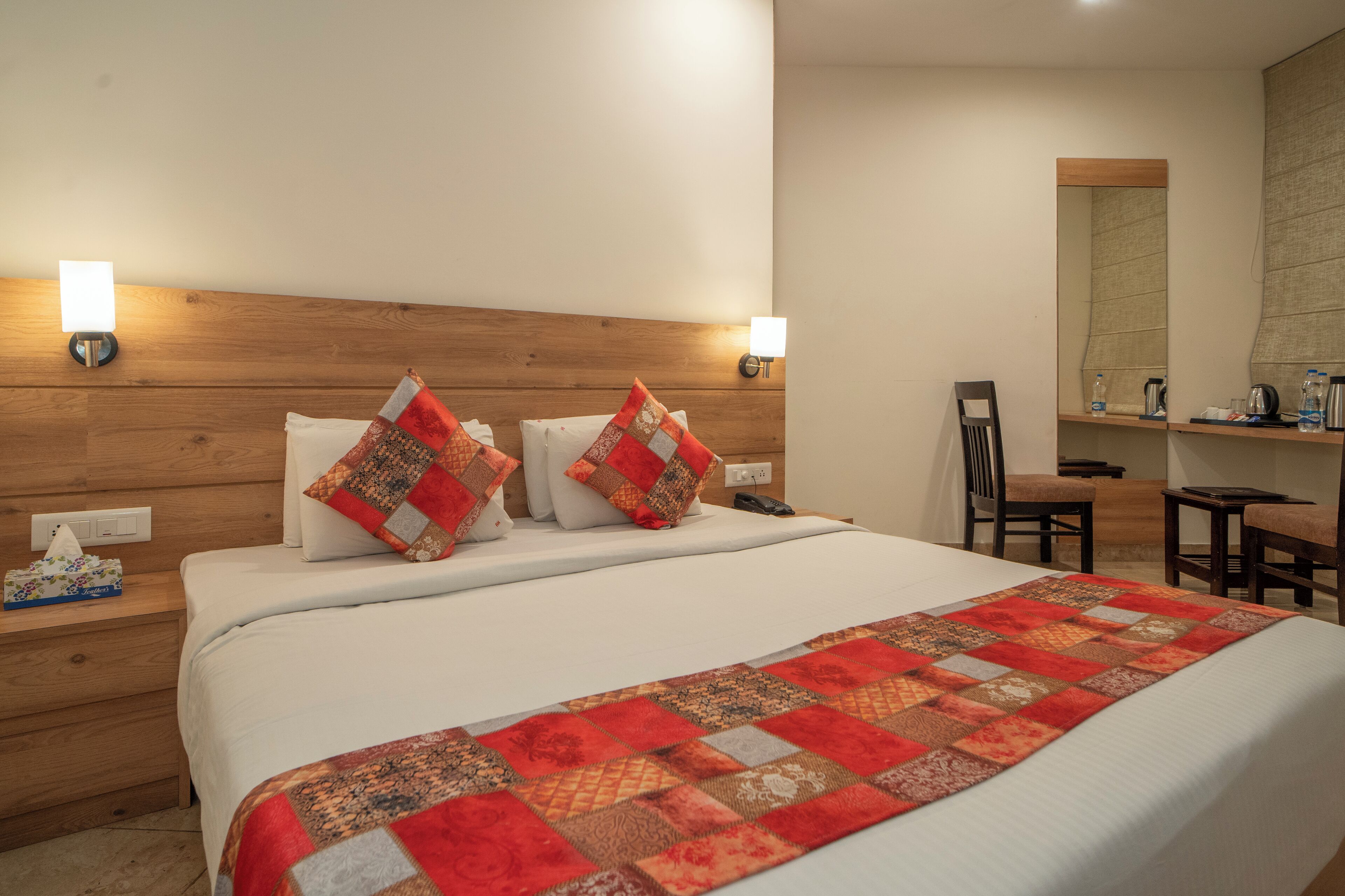 A neatly made bed with a quilt, bedside lamps, a nightstand, a chair, a mirror, and a small table with toiletries.