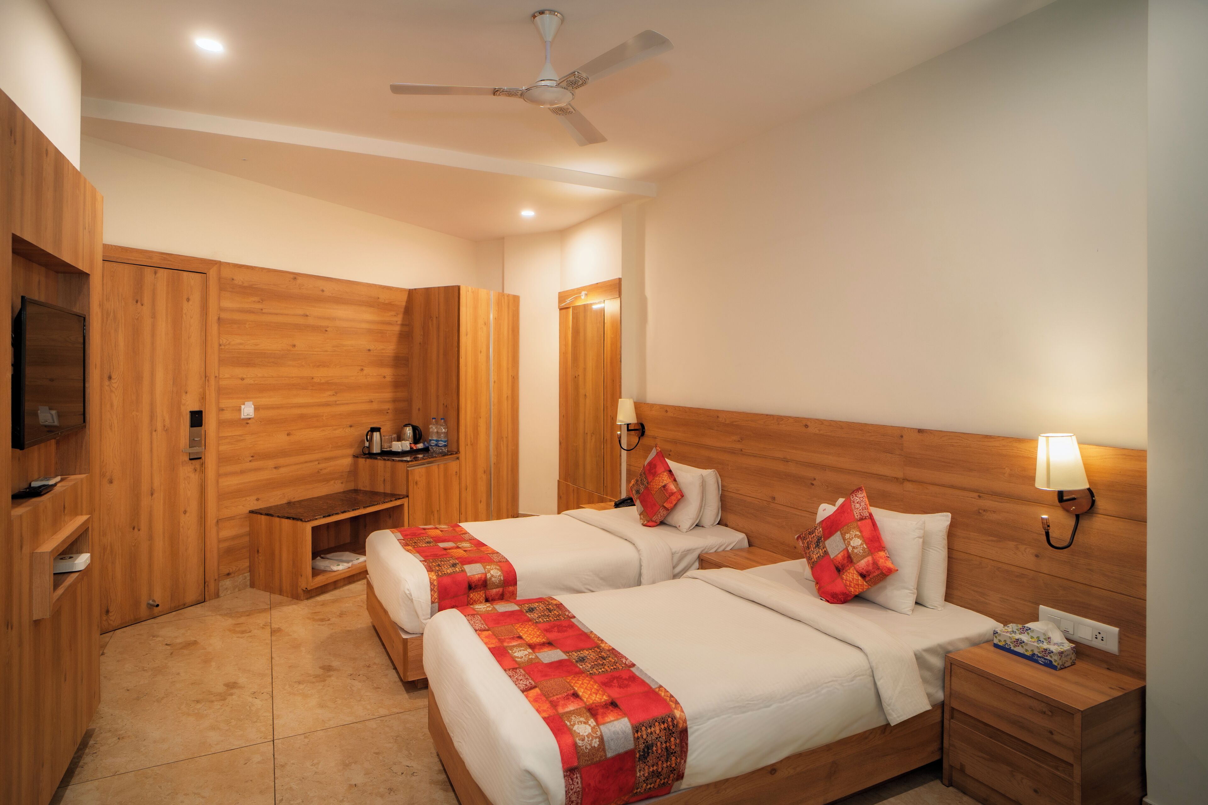 A hotel room with two beds, wooden walls, a ceiling fan, a bedside table with a lamp, and a TV on a shelf.