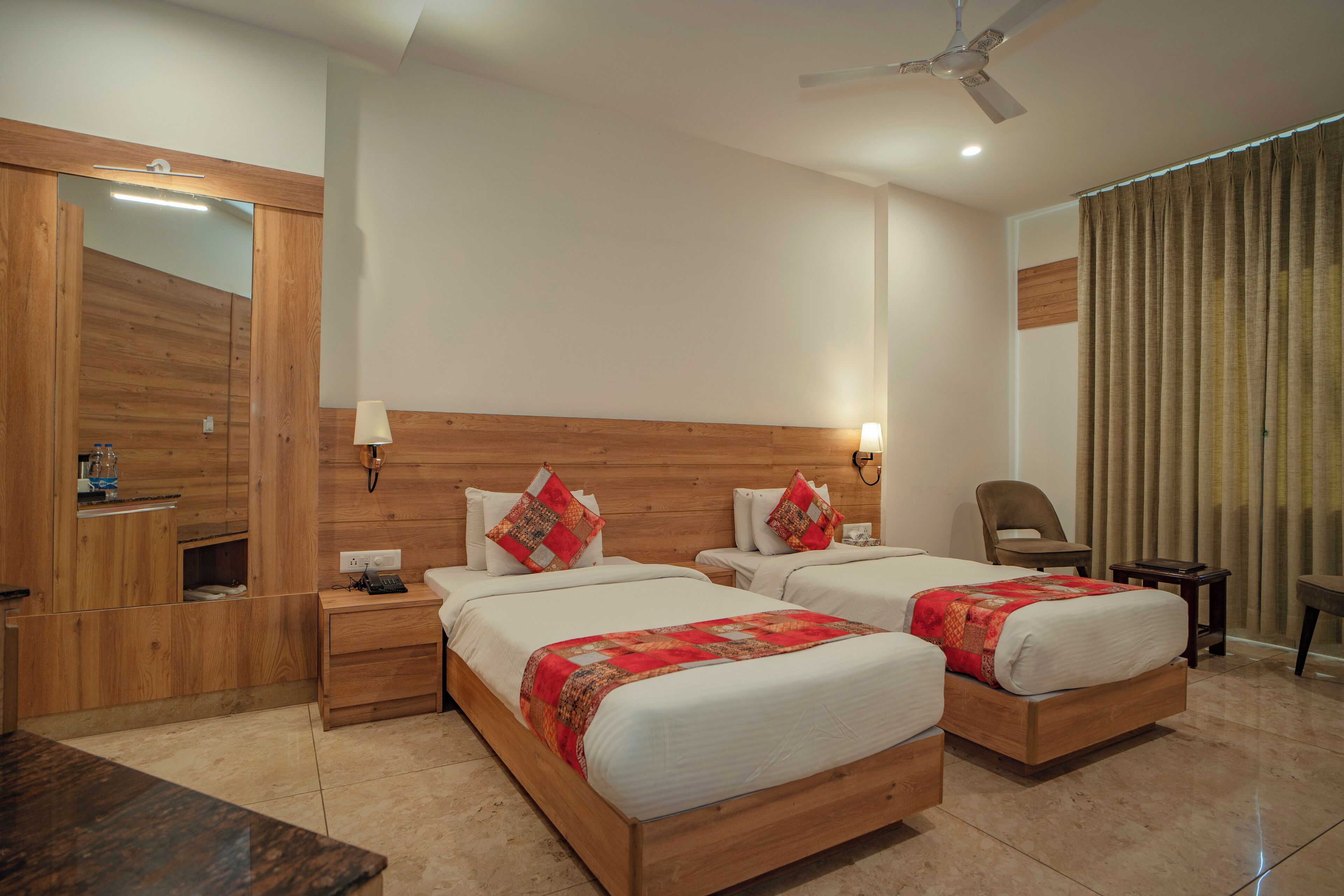 A hotel room with two beds, wooden headboard, bedside tables, a mirror, a ceiling fan, and a window with curtains.