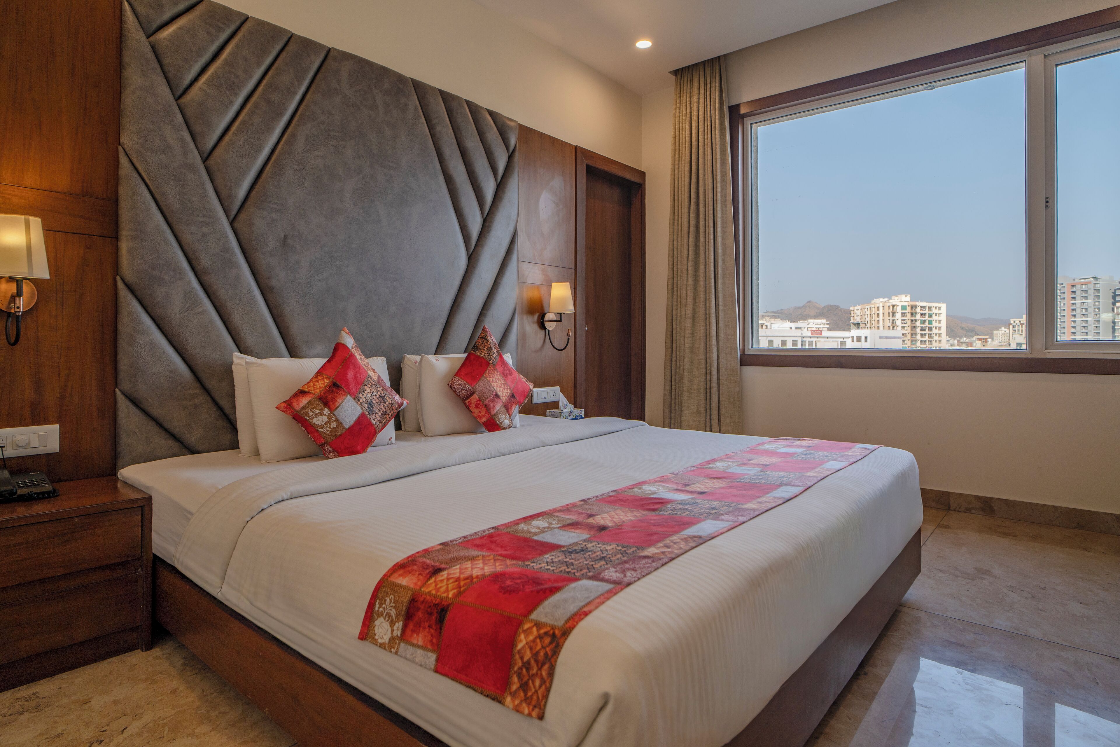 A hotel room with a large bed, a nightstand, a wall-mounted lamp, and a window with a city view.