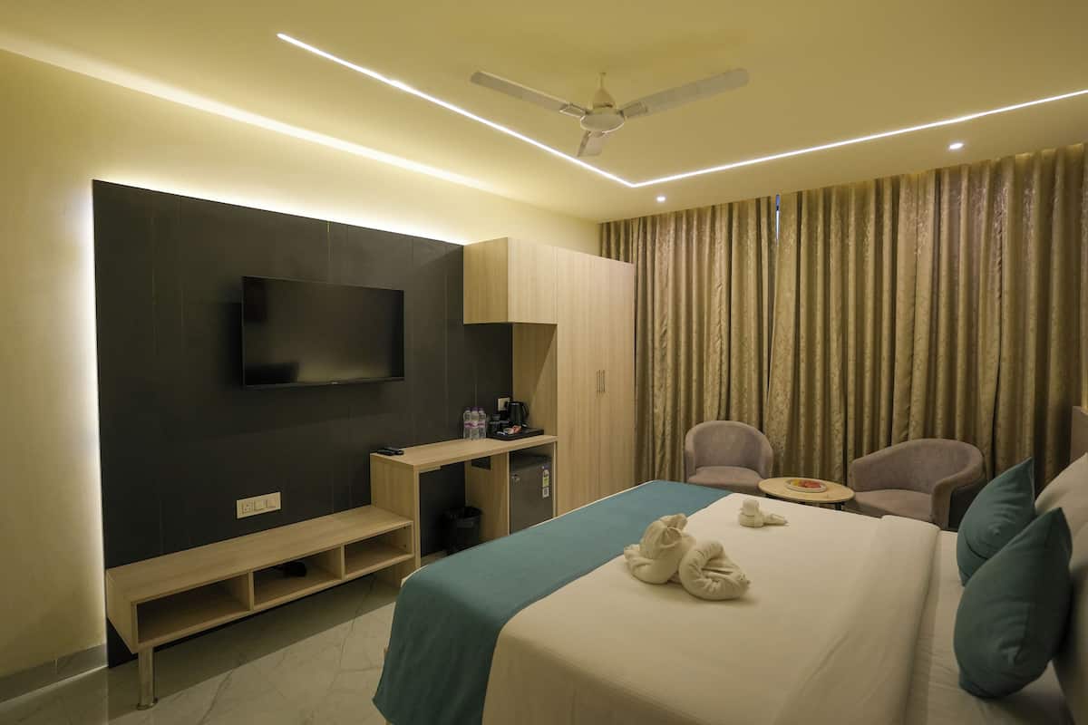A hotel room with a bed, a TV mounted on the wall, a desk with a chair, a small table, and a ceiling fan.