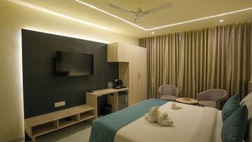 A hotel room with a bed, a TV mounted on the wall, a desk with a chair, a small table, and a ceiling fan.