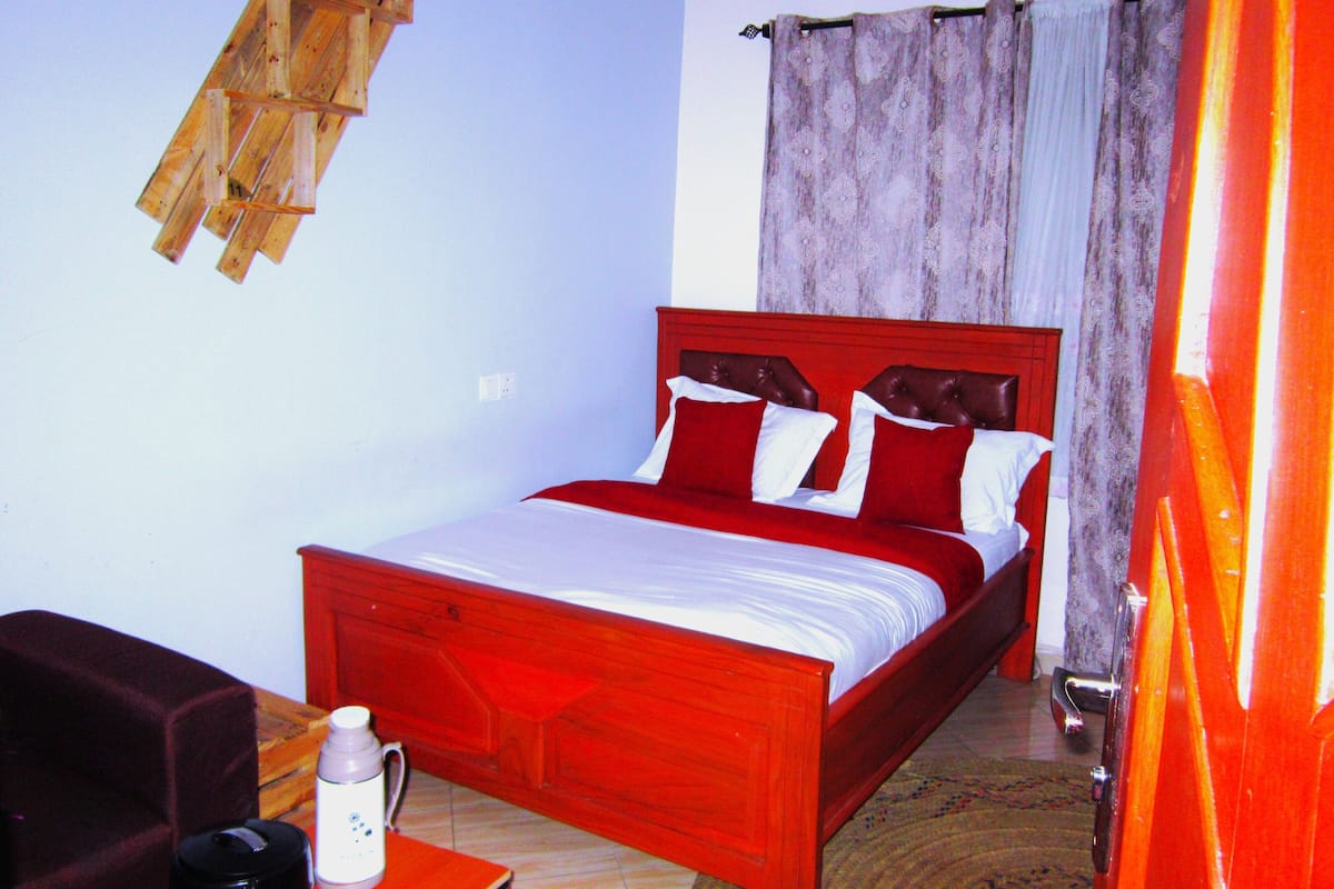 Ein Zimmer mit einem hölzernen Bett, roter Bettwäsche, einem weißen Kissen, einem Holz-Kopfteil, einer weißen Wand, einem Fenster mit Vorhängen und einem kleinen Tisch mit Wasserkocher und Tasse.