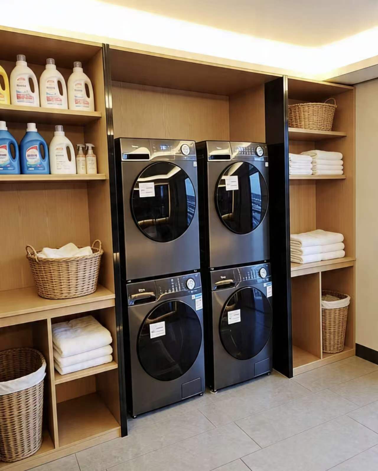 Une buanderie équipée de deux machines à laver et deux sèche-linge à chargement frontal superposés, d’étagères contenant des produits de lessive et de paniers pour le linge plié.