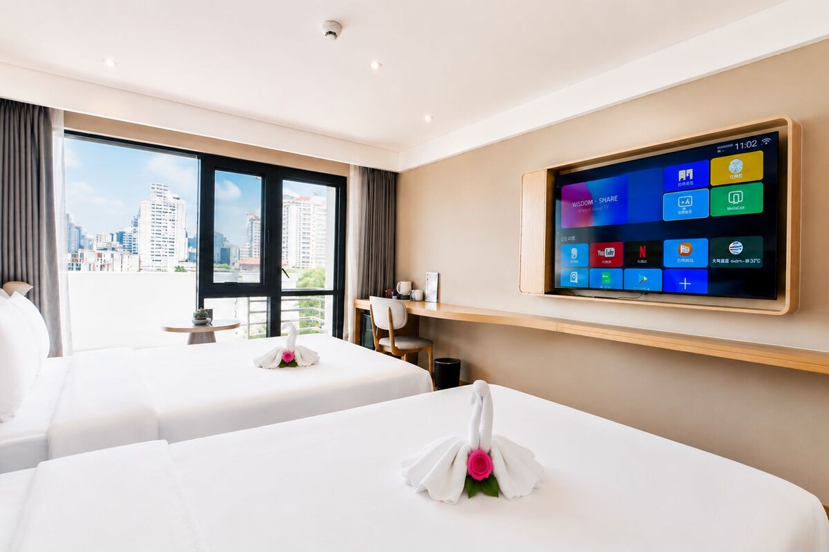 A hotel room with two beds, a flat-screen TV mounted on the wall, and a view of the city through large windows.