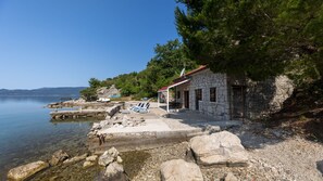 A stone building with a red roof near a calm lake, surrounded by trees and rocky terrain.