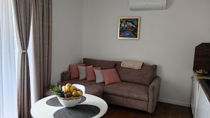 Uma sala de estar com um sofá marrom, uma mesa redonda pequena com uma fruteira, um quadro na parede e um ar condicionado branco.