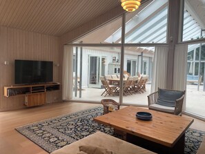 A spacious living area with a large TV, wooden furniture, a dining table, and a view through large glass doors.